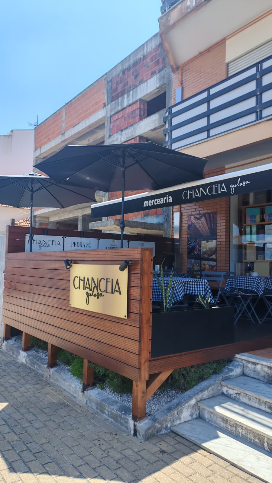 Esplanada da Mercearia Chanceia Gulosa com deck de madeira, guarda-sóis pretos e mesas xadrez.